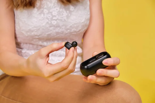 A brunette girl in summer clothes on a yellow background takes out wireless headphones. — Stock fotografie