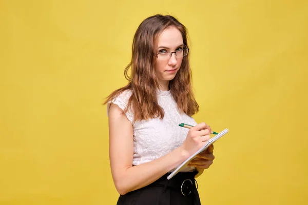 Strict teacher girl with glasses on a yellow background makes a note with a pen in a notebook. — Φωτογραφία Αρχείου