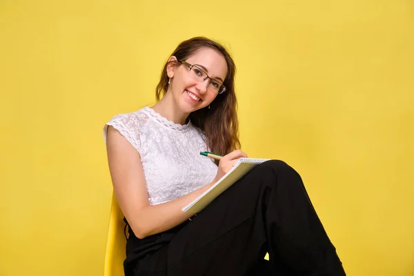 Chica sonriente profesor en gafas sobre un fondo amarillo se sienta y escribe un bolígrafo en un cuaderno . —  Fotos de Stock