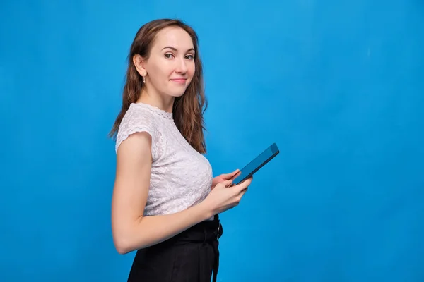 A brunette girl in casual clothes on a blue background and looks at the camera and holds a tablet. — Φωτογραφία Αρχείου