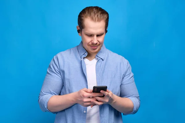 A man in a blue shirt has put on wireless headphones and is looking at the smartphone while talking. — ストック写真