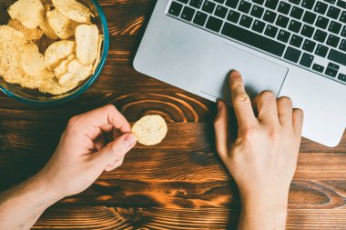 Glass plate with chips next to a laptop on a burnt wooden background. Man has chips in his hands and browses the Internet. Toned. Top view.