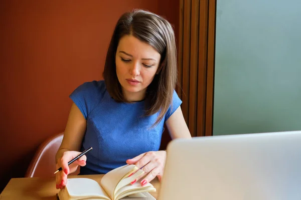 The girl works at a laptop and looks at the notebook. The concept of the schedule of meetings for the day.