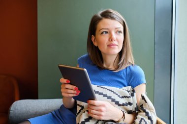 A girl in a blue dress sits at a cafe with a tablet and dreams.