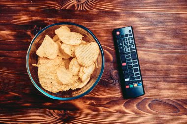 Chips in a glass dish next to the TV remote control against a burnt wooden background. Top view. Toned.