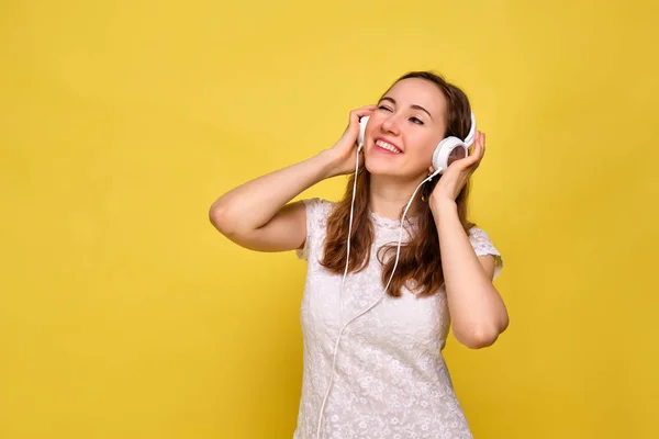 Девушка в футболке на жёлтом фоне слушает музыку в белых наушниках и подмигивает. Концепция платной подписки для прослушивания аудио . — стоковое фото