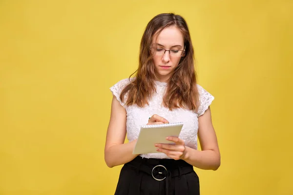 A girl in casual clothes and glasses on a yellow background makes notes in a notebook. Business woman concept. — Stock fotografie