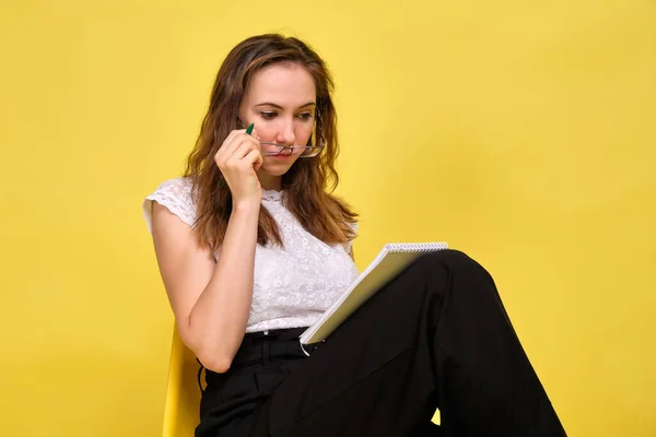 A girl in casual clothes and glasses on a yellow background sits makes a plan for the day in a notebook. The concept of the distribution of the day. — Stock fotografie