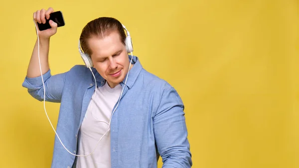 Un homme en chemise bleue sur fond jaune écoute de la musique dans des écouteurs blancs et danse. Espace de copie . — Photo
