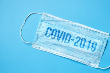 Mavi arkaplanda tıbbi yüz maskesi, Covid-2019 yazısıyla. Yaklaş..