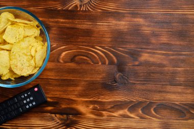 Patates cipsi, uzaktan kumandalı televizyon kumandasının yanındaki cam kapta fotokopi alanı olan ahşap bir arka planda. Üst Manzara. Kapat..