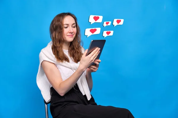 一个穿着蓝色背景休闲装的女孩坐在社交网络上 看着平板电脑 以图标的形式接收抽象的喜欢 靠近点 — 图库照片