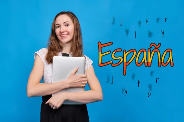 A girl in casual clothes on a blue background holds a tablet that is learning Spanish. Abstract word for spain in spanish, country colors. Close up.