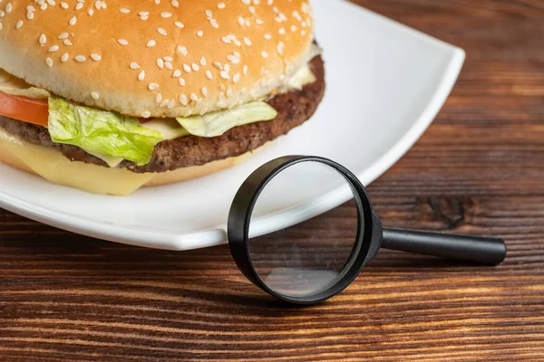 Burger on a white plate and a wooden background with a magnifier. Composition research concept. Close up.