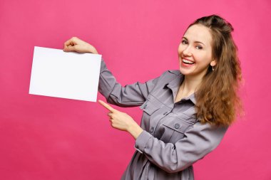 Pembe arka planda gri elbiseli beyaz bir kız. Beyaz bir kağıt parçası tutuyor ve gülümsüyor. Uzayı kopyala Kapat..