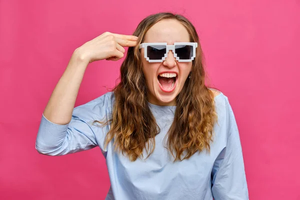 Stylish caucasian girl in a pale blue t-shirt and 8-bit glasses and an abstract gun from his hand pushed to her head smiling on a pink background. Close up.