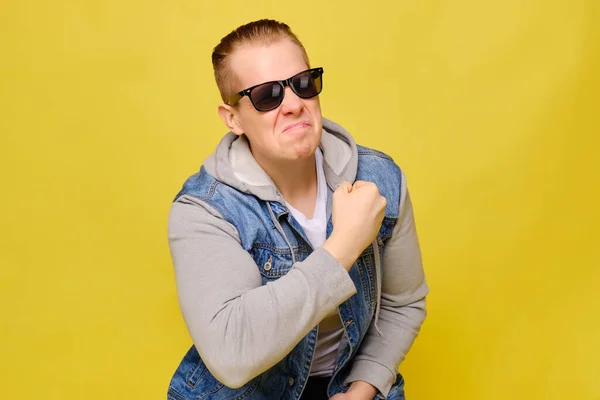 Stylish Caucasian man in a jeans and sunglasses on a yellow background. Close up. Jokingly demonstrates biceps arms.