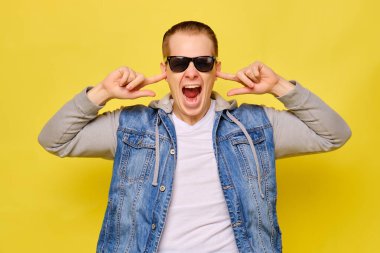 Sarı arka planda kot pantolon ve güneş gözlüğü takan şık beyaz bir adam. Kapatın. Elinin parmaklarıyla kulaklarını kapatıyor..