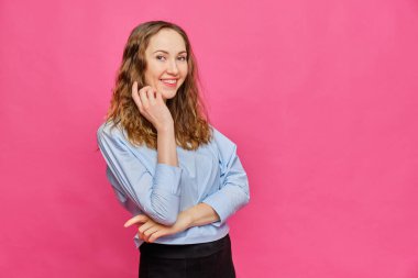 Soluk mavi tişörtlü beyaz kız pembe arka planda kameraya şakacı bir şekilde bakıyor. Kapat..