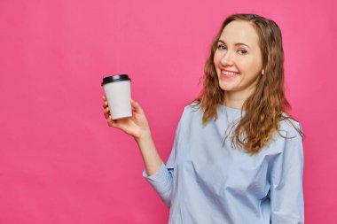 Pembe arka planda kahve için kağıt bardak gösteren açık mavi tişörtlü beyaz bir kız. Uzayı kopyala Kapat..