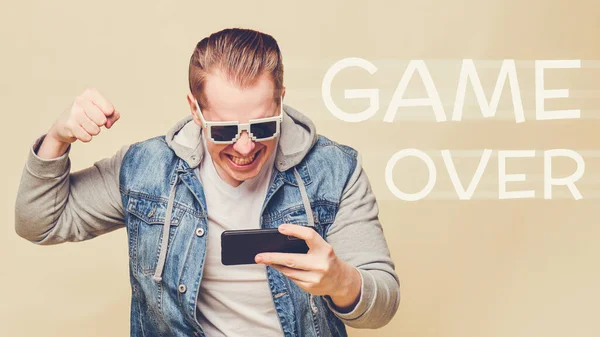 黄色の背景に8ビットのメガネをかけたジーンズを着たスタイリッシュな白人男性 スマートフォンで遊ぶことと負けることのコンセプトはゲームオーバーです 閉じろ — ストック写真