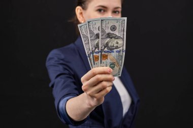 Ceketli kız, siyah arka planda kameraya yönelttiğimiz dolarları elinde tutuyor. Kapat..