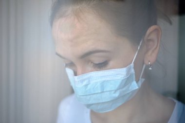 Tıbbi maskeli kız hastanede karantinaya alındı ve alnını pencereye dayadı. Kapat..