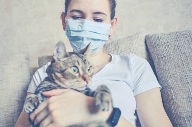 Tıbbi maskeli bir kız evde karantinada, kollarında bir kedi tutuyor. Ton halinde. Kapat..