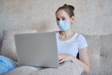 Tıbbi maskeli bir kız evde karantinada dizüstü bilgisayarla çalışıyor. Kapat..