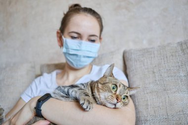 Tıbbi maskeli kız evde kendini izole ediyor ve kediye bakıyor. Kapat..