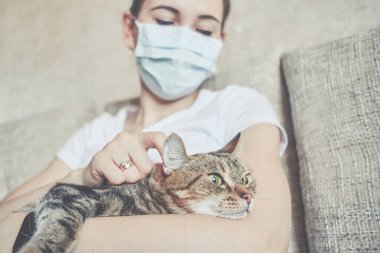 Tıbbi maskeli kız evde kendini izole ediyor ve bir kediyle kanepede dinleniyor. Kapat..