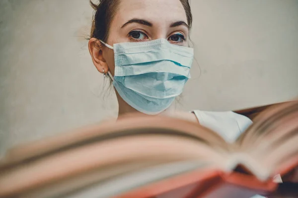 The girl in a medical mask is on self-isolation at home holding a book and looking at the camera. Close up.