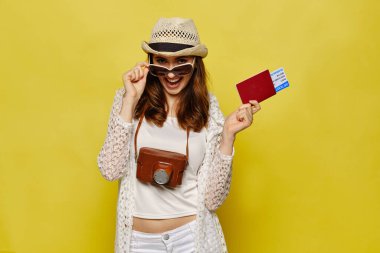 Şapkalı, uçak bileti olan, pasaportlu bir kız sarı arka plandaki kameraya bakıyor. Kapat..