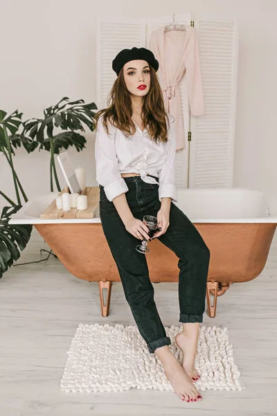 Young cute woman in black beret and red lips drinking a glass of wine and seating on bath-tub — Stock Photo, Image