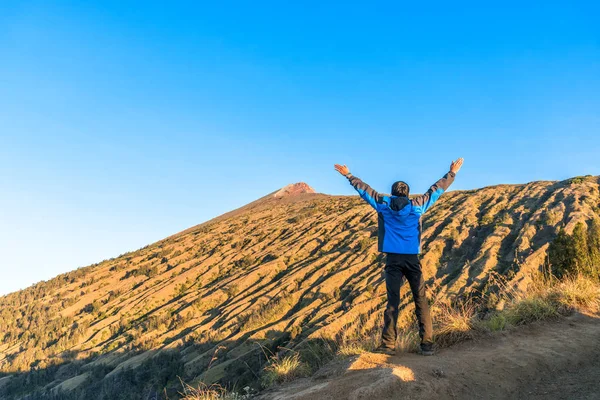 Jeune homme randonneur porter une veste bleue, écarter la main, profiter et heureux avec vue sur le sommet de la montagne après avoir terminé l'escalade à la montagne Rinjani, Lombok, Indonésie . — Photo