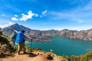 Hiker man with backpack spreading hand, enjoy and happy with active volcano Baru Jari, Lake Segara Anak and summit of Rinjani mountain view after finished climbing at Rinjani mountain. clipart