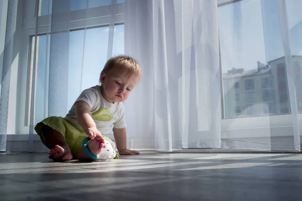 Słodkie dziecko w domu, w białym pokoju siedzi w pobliżu okna. Piękne dziecko może być chłopak lub dziewczyna i ma na sobie garnitur ciała. Baby z kręconych usłyszeć, to patrząc na kamery. Zdjęcia Stockowe bez tantiem