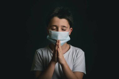 Coronavirus için tıbbi maske takan, elleri birbirine kenetlenmiş ve siyah üzerine dua eden bir çocuk.