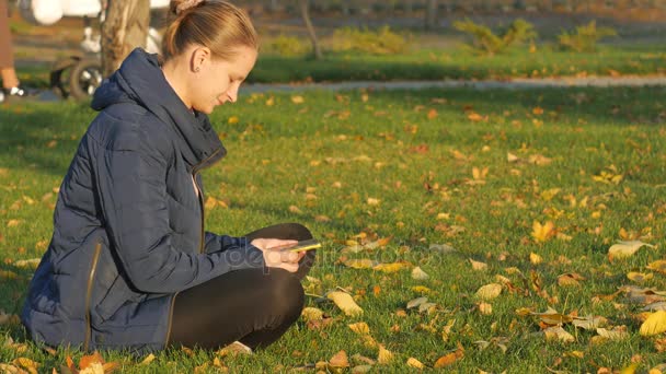 Outono menina no parque senta-se na grama, enviando sms do telefone . — Vídeo de Stock