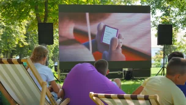 Ucraina, Kirovograd luglio 2019. Un sacco di gente si siede sull'erba in un parco pubblico e guarda un film, cartone animato. Street Festival. Mostrare un film all'aperto. Bambini in strada evento . — Video Stock