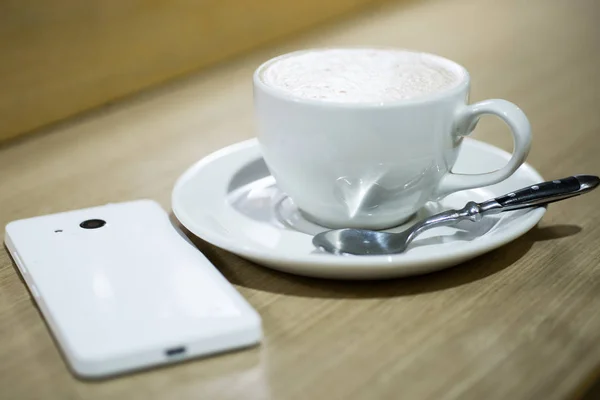 Kawa cappuccino w białe filiżanka i biały telefon na stole w kawiarni. — Zdjęcie stockowe