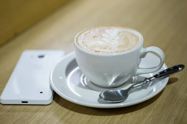 Καφές καπουτσίνο στο φλιτζάνι λευκό και λευκή κινητό τηλέφωνο στο τραπέζι σε μια καφετέρια. — Φωτογραφία Αρχείου
