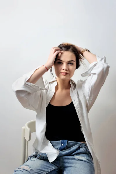 Modelo de moda atraente posando na cadeira em jeans e camisa segurar o cabelo mãos — Fotografia de Stock