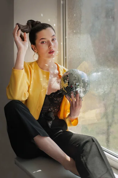 Young woman with bright colorfull spice makeup and Mirror disco ball with shines on shes body near window — Stockfoto