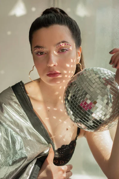 Young woman with bright colorfull spice makeup and Mirror disco ball with shines on shes body near window — Stockfoto