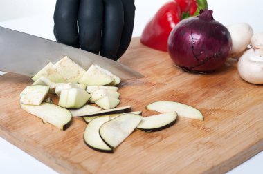 Siyah eldivenli şef bambu tahtasında patlıcan dilimliyor.