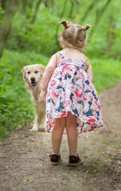 Orman yolunda duran köpekli, açık renk elbiseli mutlu küçük kız. Şirin küçük kız köpekle oynuyor, evcil hayvanlar günü.