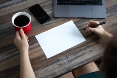 Eller boş kağıda kahve fincanı, telefon, dizüstü bilgisayar ve ahşap masa üzerine fotokopi alanı ile yazılıyor. Üst görünüm, kapat