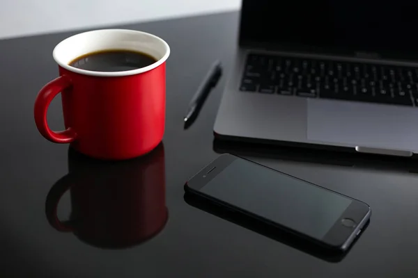 Stylish red coffee cup, phone and laptop on desk table. Black background, business concept, close up