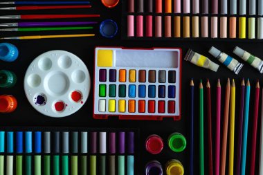Colorful stationery flat lay: pastel crayons, colored pencils, watercolors on black background. Art workspace school concept. Top view clipart